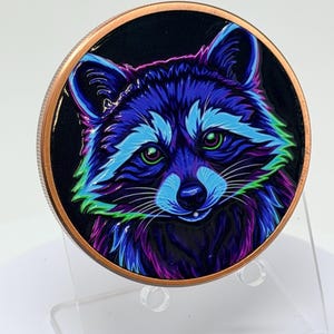 May include: A circular copper-toned coin featuring a vibrant, stylized illustration of a raccoon. The raccoon is rendered in neon shades of blue, purple, and green against a solid black background, with bright green eyes and detailed fur.