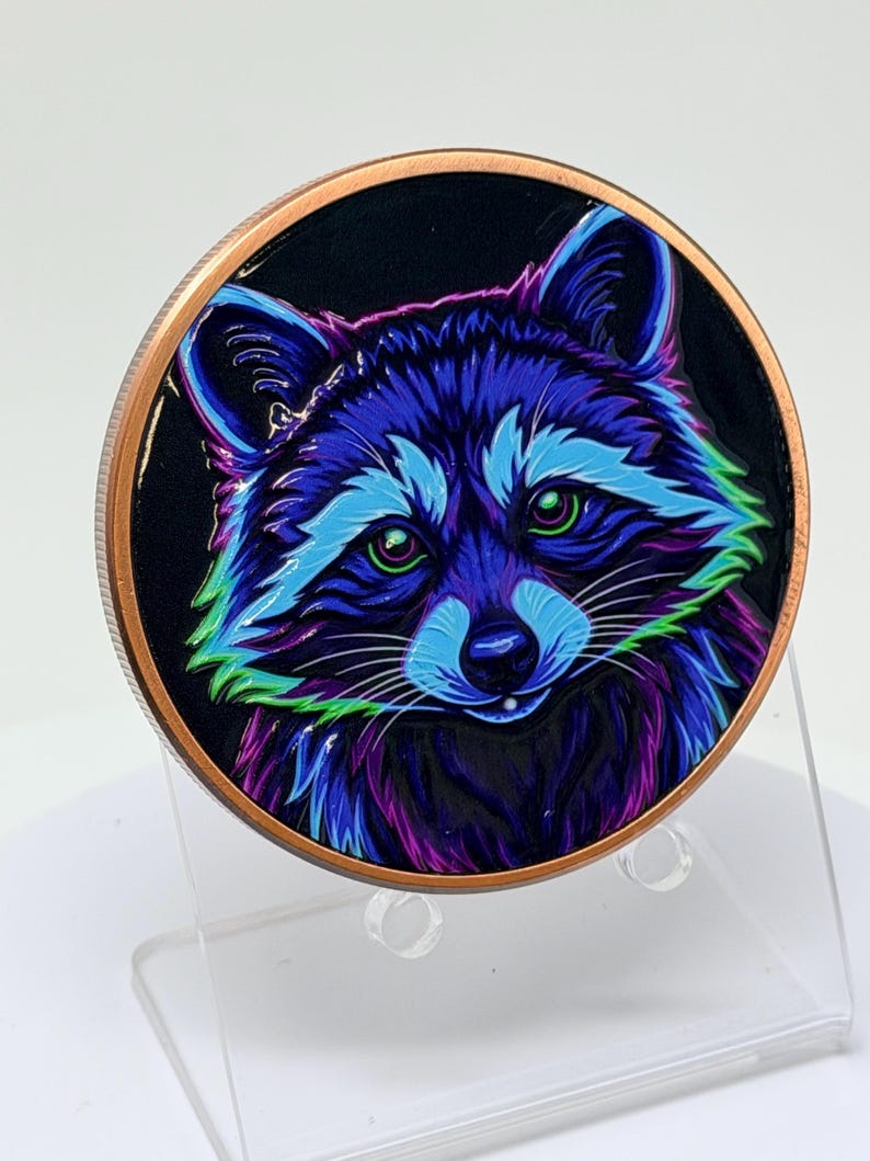 May include: A circular copper-toned coin featuring a vibrant, stylized illustration of a raccoon. The raccoon is rendered in neon shades of blue, purple, and green against a solid black background, with bright green eyes and detailed fur.