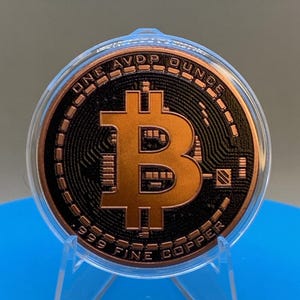 A copper-colored Bitcoin round featuring a central Bitcoin symbol in relief against a black textured background. The text ONE AVDP OUNCE is inscribed at the top, and .999 FINE COPPER is inscribed at the bottom. It is encased in a clear plastic protective capsule and displayed in a stand.