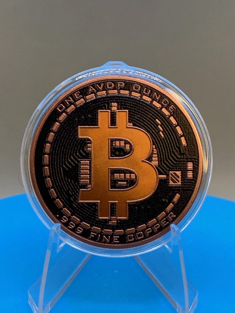 A copper-colored Bitcoin round featuring a central Bitcoin symbol in relief against a black textured background. The text ONE AVDP OUNCE is inscribed at the top, and .999 FINE COPPER is inscribed at the bottom. It is encased in a clear plastic protective capsule and displayed in a stand.