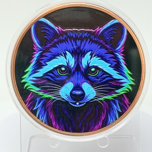 May include: A circular medallion featuring a vibrant, neon-colored illustration of a raccoon face. The raccoon is rendered in shades of electric blue, bright purple, and lime green against a black background, encased in a clear, round frame.