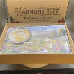 Wider view of the open Harmony Bee copper round packaging box on a table, with the round in its capsule resting on the printed certificate insert.