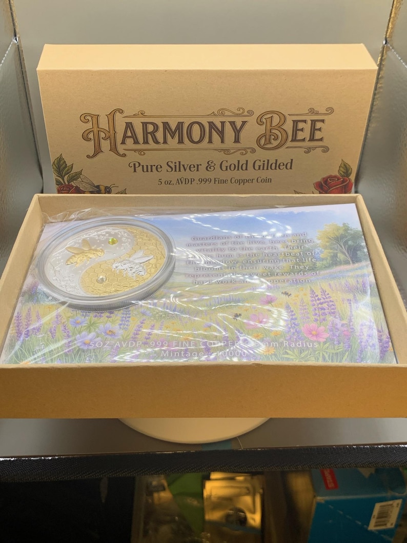 Wider view of the open Harmony Bee copper round packaging box on a table, with the round in its capsule resting on the printed certificate insert.