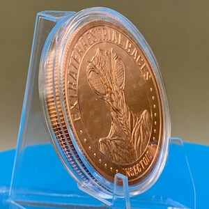 Tilted side view of the Intaglio Mint Insectoid 1 oz copper round in its clear capsule on a stand, showing the thickness and raised rim of the round and the EXTRATERRESTRIAL RACES obverse design.