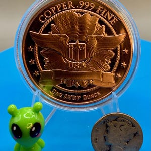 Reverse of a 1 oz .999 fine copper round in a display holder next to a small green alien figurine and a worn silver dime for size comparison, on a blue background.