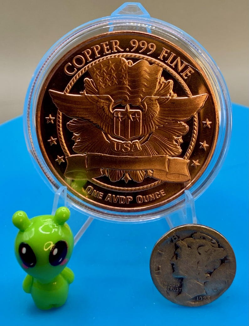 Reverse of a 1 oz .999 fine copper round in a display holder next to a small green alien figurine and a worn silver dime for size comparison, on a blue background.