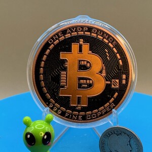 A copper-colored Bitcoin round in a clear plastic case, featuring the text ONE AVDP OUNCE and 999 FINE COPPER. Beside it sits a small green alien figurine and a silver-colored United States coin dated 1906 with a profile portrait.