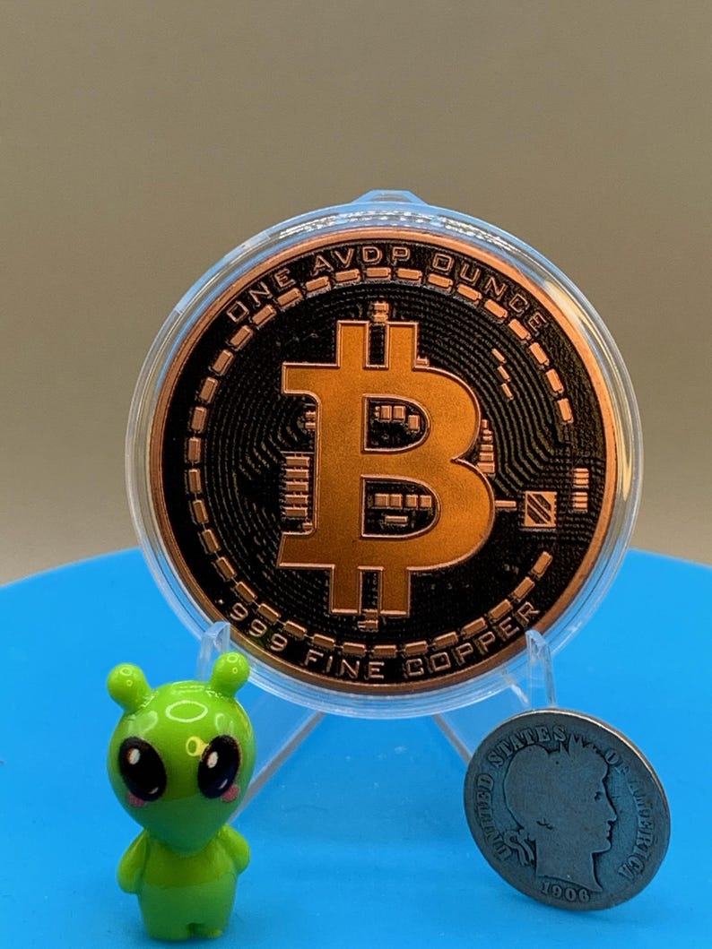 A copper-colored Bitcoin round in a clear plastic case, featuring the text ONE AVDP OUNCE and 999 FINE COPPER. Beside it sits a small green alien figurine and a silver-colored United States coin dated 1906 with a profile portrait.