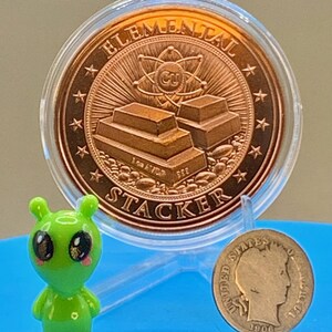 "Elemental Stacker" reverse of the First Contact 1 oz copper round in its capsule next to a small green alien figurine and a silver coin for size comparison