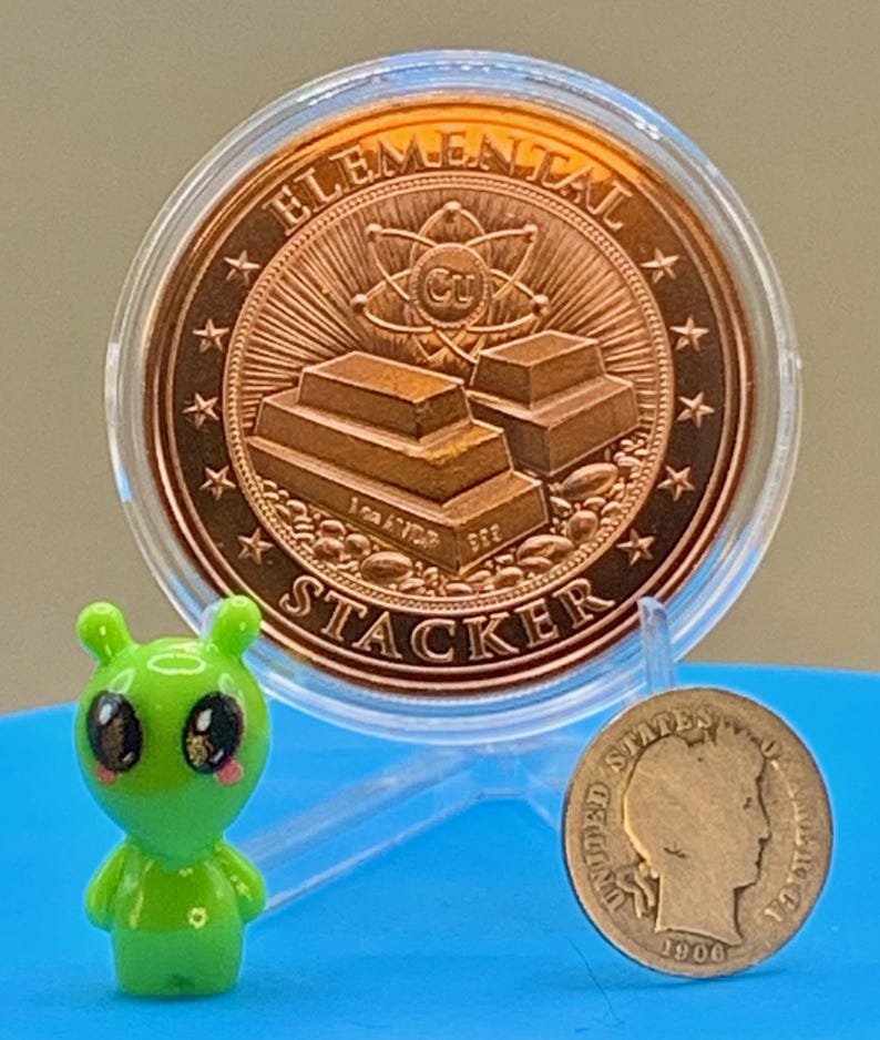 "Elemental Stacker" reverse of the First Contact 1 oz copper round in its capsule next to a small green alien figurine and a silver coin for size comparison
