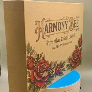 Angled view of the Harmony Bee copper round's kraft-paper packaging box standing upright next to its round display case.