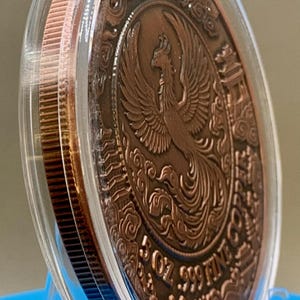Angled side view of the copper Phoenix round in its protective capsule, showing the coin's edge and thickness against a blue background.