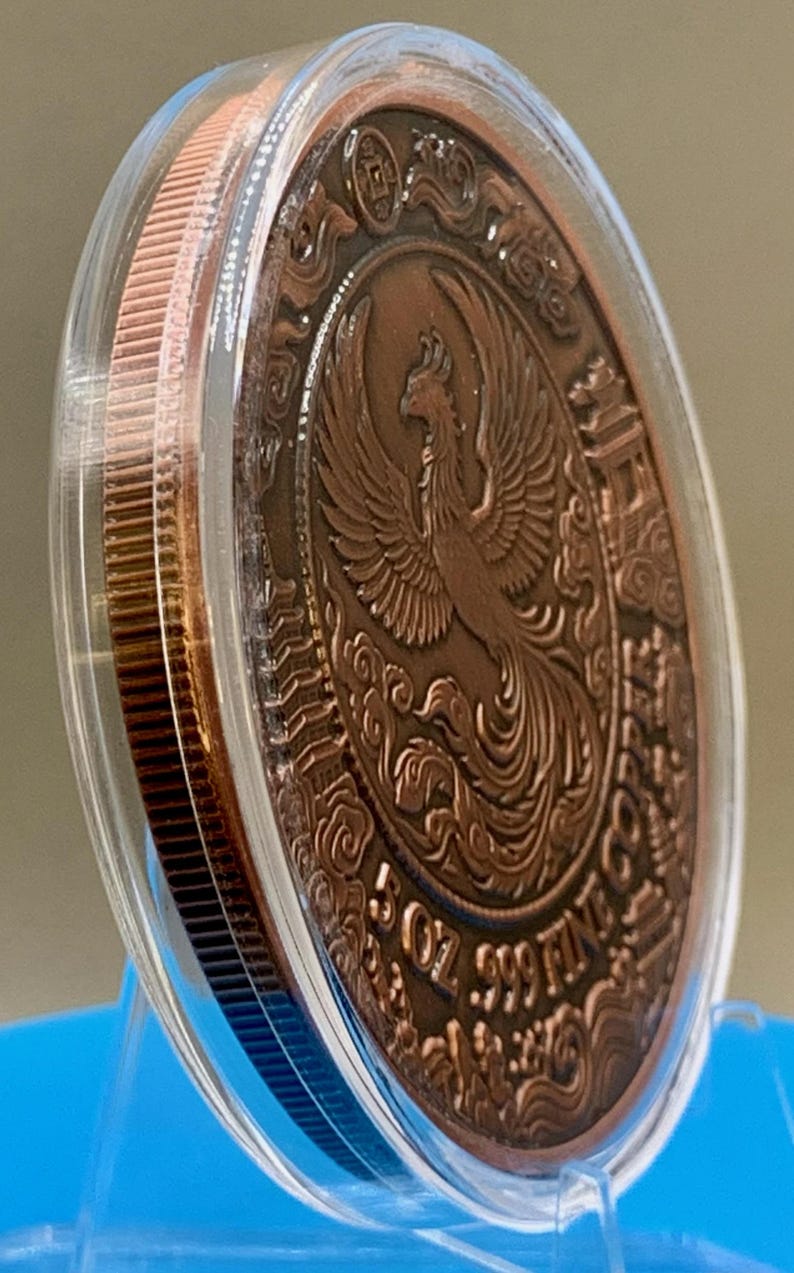Angled side view of the copper Phoenix round in its protective capsule, showing the coin's edge and thickness against a blue background.