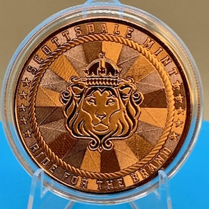 Peut inclure: Une pi&egrave;ce de monnaie ronde de couleur cuivre enferm&eacute;e dans une capsule de protection en plastique transparent. La pi&egrave;ce pr&eacute;sente une t&ecirc;te de lion couronn&eacute;e au centre, entour&eacute;e d'une bordure en corde, du texte SCOTTSDALE MINT et RIDE FOR THE BRAND.