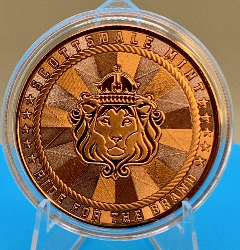Peut inclure: Une pi&egrave;ce de monnaie ronde de couleur cuivre enferm&eacute;e dans une capsule de protection en plastique transparent. La pi&egrave;ce pr&eacute;sente une t&ecirc;te de lion couronn&eacute;e au centre, entour&eacute;e d'une bordure en corde, du texte SCOTTSDALE MINT et RIDE FOR THE BRAND.