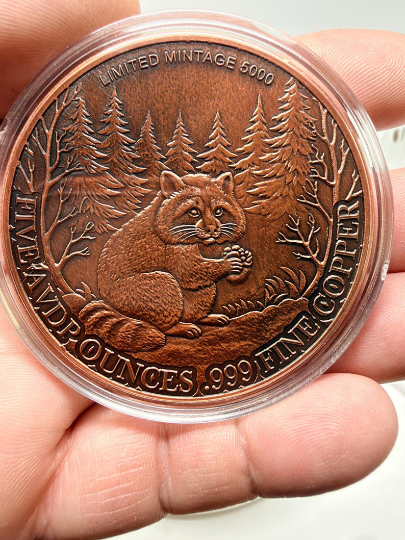 May include: A round copper coin featuring a relief of a raccoon holding a pinecone in a forest setting. The text reads LIMITED MINTAGE 5000, FIVE AVD P OUNCES, and .999 FINE COPPER. The coin is encased in a clear protective plastic capsule.