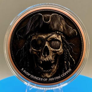 Puede incluir: Una moneda de cobre redonda que presenta un cr&aacute;neo detallado con un sombrero de pirata y un parche en el ojo. La moneda tiene un acabado oscuro y desgastado, con el texto 5 AVDP OUNCES OF .999 FINE COPPER grabado en el borde inferior.