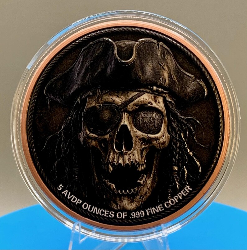 Puede incluir: Una moneda de cobre redonda que presenta un cr&aacute;neo detallado con un sombrero de pirata y un parche en el ojo. La moneda tiene un acabado oscuro y desgastado, con el texto 5 AVDP OUNCES OF .999 FINE COPPER grabado en el borde inferior.