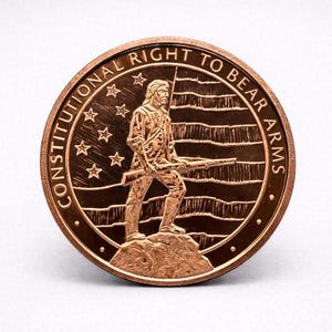 May include: A bronze-colored coin with the text "CONSTITUTIONAL RIGHT TO BEAR ARMS" encircling the edge. The coin features a detailed depiction of a person holding a rifle, standing on a rock, with a flag in the background and stars.