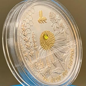 Angled close-up of the reverse side of the Harmony Bee copper round in its capsule, showing light reflecting off the raised gold bee and coneflower engraving.
