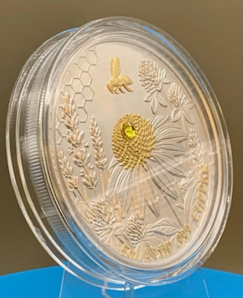 Angled close-up of the reverse side of the Harmony Bee copper round in its capsule, showing light reflecting off the raised gold bee and coneflower engraving.