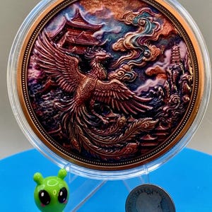 Copper Phoenix round with an iridescent patina finish in its capsule, shown next to a small green alien figurine and a dime for size comparison, Area51 Precious Metals themed display.