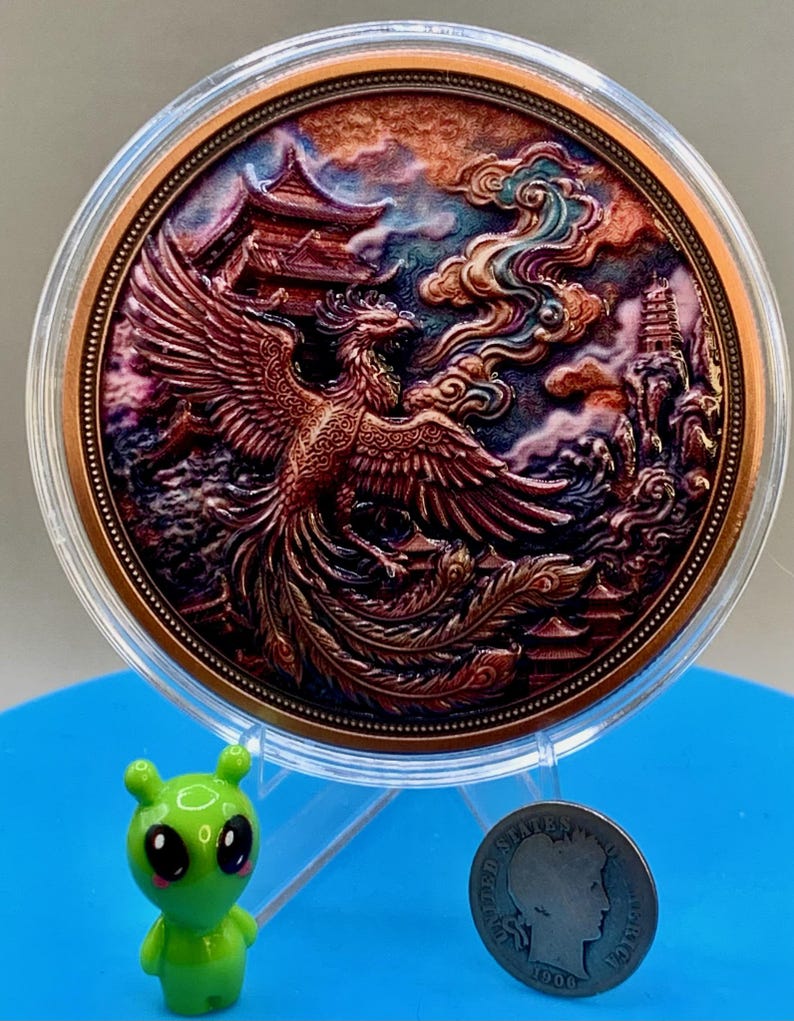 Copper Phoenix round with an iridescent patina finish in its capsule, shown next to a small green alien figurine and a dime for size comparison, Area51 Precious Metals themed display.