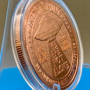 Tilted side view of the Intaglio Mint UFO Sightings 1 oz copper round in its clear capsule on a stand, showing the coin's edge and the reverse flying saucer and tractor beam design.