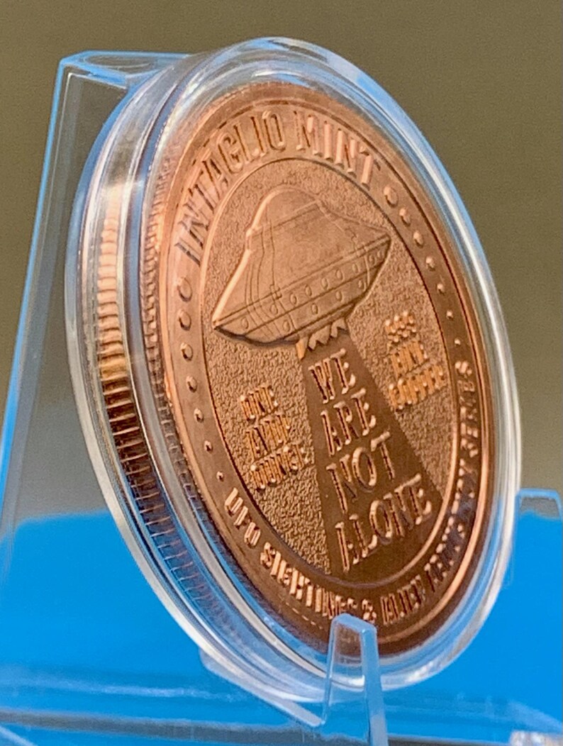 Tilted side view of the Intaglio Mint UFO Sightings 1 oz copper round in its clear capsule on a stand, showing the coin's edge and the reverse flying saucer and tractor beam design.