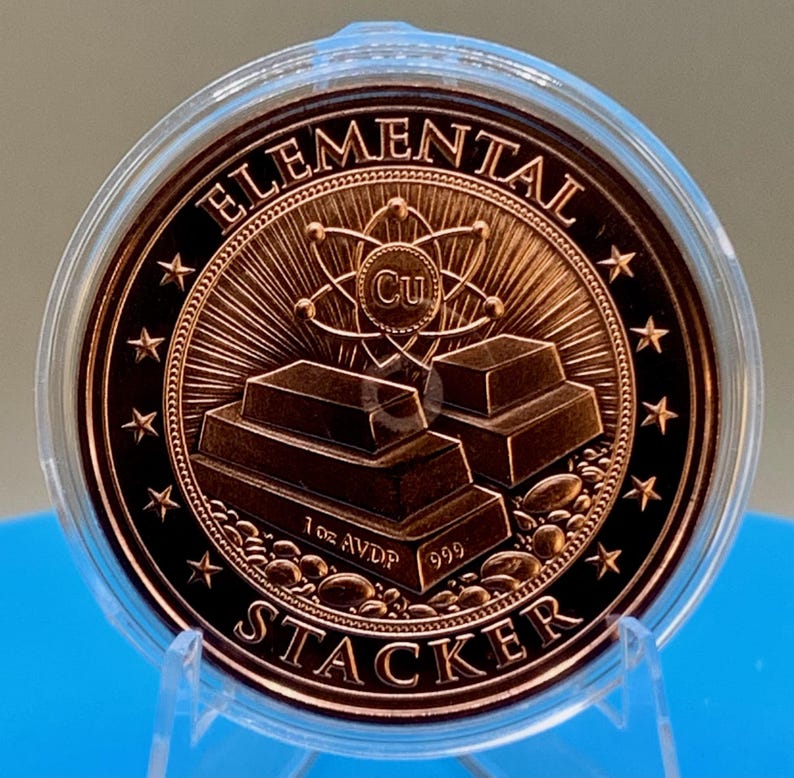 Close-up of the reverse of the 1 oz .999 fine copper round in a clear capsule, showing stacked copper bars with an atom and the Cu symbol, "Elemental" across the top and "Stacker" along the bottom, surrounded by a ring of stars.