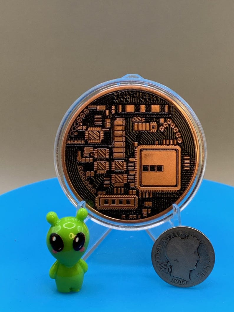 A circular copper-colored Bitcoin round featuring a black circuit board pattern, encased in a clear plastic protective holder. Beside it stands a small, bright green alien figurine and a silver-colored 1908 United States Barber dime.