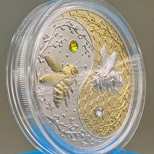 Close-up of the Harmony Bee copper round inside its clear protective capsule, angled view showing the gold and silver gilded bee design and honeycomb texture.