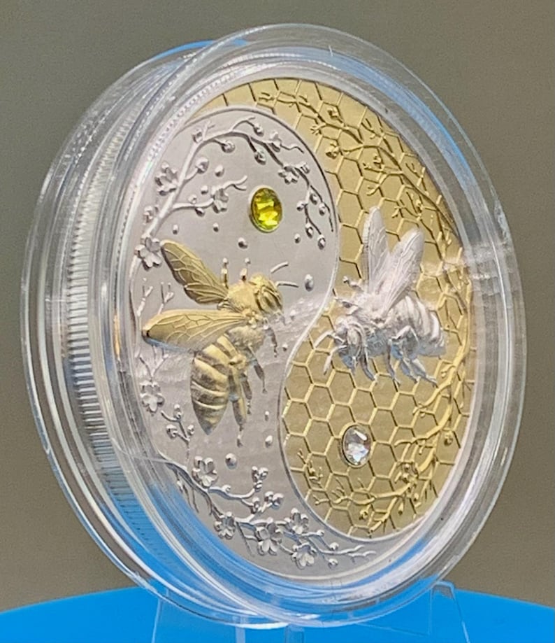 Close-up of the Harmony Bee copper round inside its clear protective capsule, angled view showing the gold and silver gilded bee design and honeycomb texture.