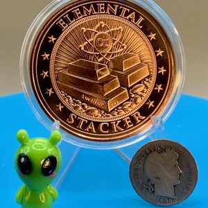 Reverse of the 1 oz copper round in a clear capsule, showing stacked copper bars with an atom and the Cu symbol and the words "Elemental" and "Stacker" around the rim, beside a small green alien figurine and an older silver-colored coin for scale.