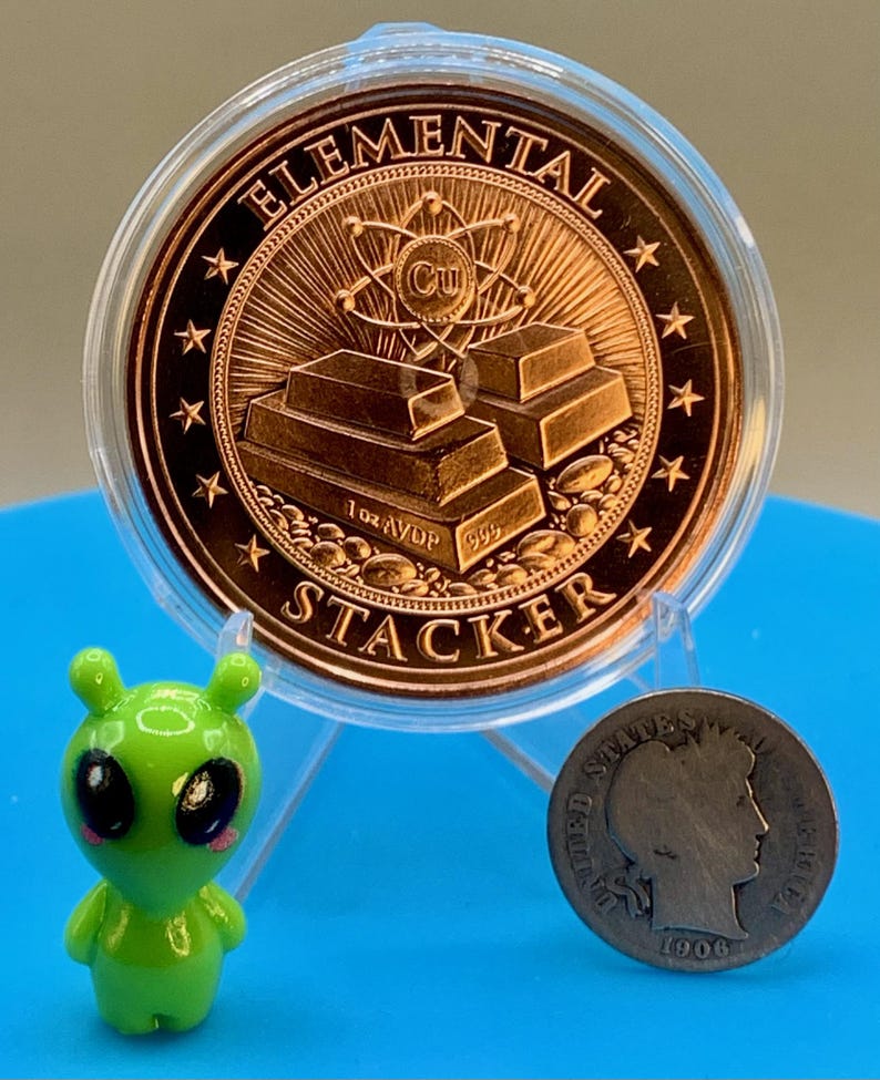 Reverse of the 1 oz copper round in a clear capsule, showing stacked copper bars with an atom and the Cu symbol and the words "Elemental" and "Stacker" around the rim, beside a small green alien figurine and an older silver-colored coin for scale.