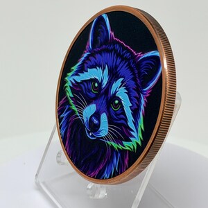 May include: A round copper-toned coin featuring a vibrant, neon-colored illustration of a raccoon against a black background. The raccoon is depicted with bright blue, purple, and green fur highlights and glowing green eyes, set on a clear stand.