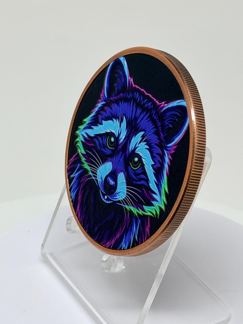 May include: A round copper-toned coin featuring a vibrant, neon-colored illustration of a raccoon against a black background. The raccoon is depicted with bright blue, purple, and green fur highlights and glowing green eyes, set on a clear stand.