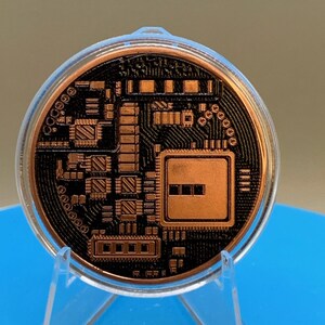 The reverse side of the copper-colored Bitcoin round, featuring a detailed black relief design of a computer circuit board with microchips, processors, and intricate wiring patterns, encased in a clear protective plastic capsule on a stand.