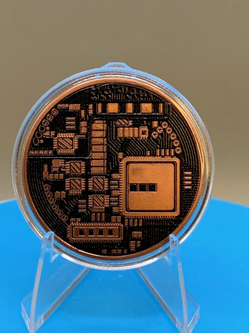 The reverse side of the copper-colored Bitcoin round, featuring a detailed black relief design of a computer circuit board with microchips, processors, and intricate wiring patterns, encased in a clear protective plastic capsule on a stand.