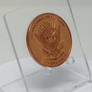 Angled rear view of the 1 oz copper round showing the Great Seal eagle reverse, tilted on a clear acrylic stand