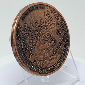 May include: A round copper coin featuring a relief of a raccoon in a forest setting. The text reads LIMITED MINTAGE 5000 and FIVE AVD P OUNCES .999 FINE COPPER. The coin has a reeded edge and is displayed on a clear plastic stand.