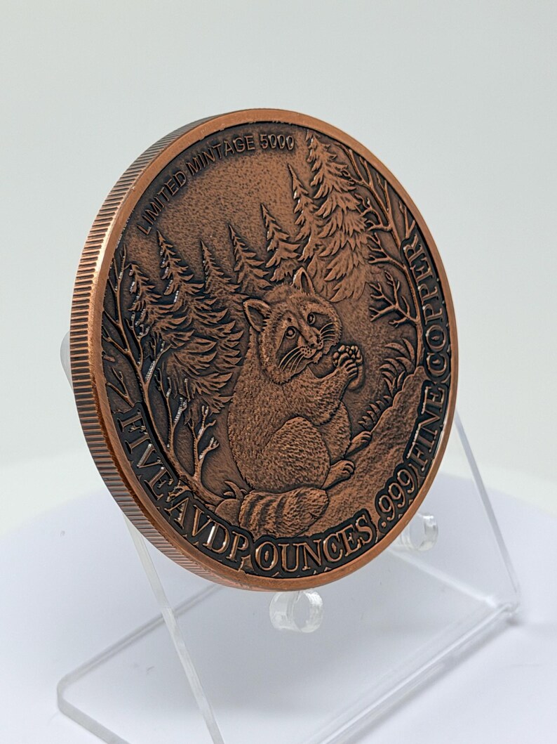 May include: A round copper coin featuring a relief of a raccoon in a forest setting. The text reads LIMITED MINTAGE 5000 and FIVE AVD P OUNCES .999 FINE COPPER. The coin has a reeded edge and is displayed on a clear plastic stand.