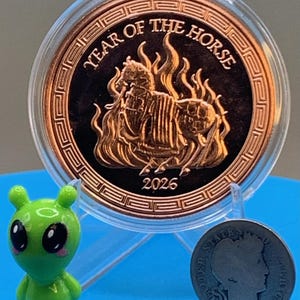 Encapsulated 2026 Year of the Horse copper round on a blue stand, armored horse obverse showing, flanked by the Area 51 Precious Metals green alien mascot figurine and a silver Barber dime for scale.