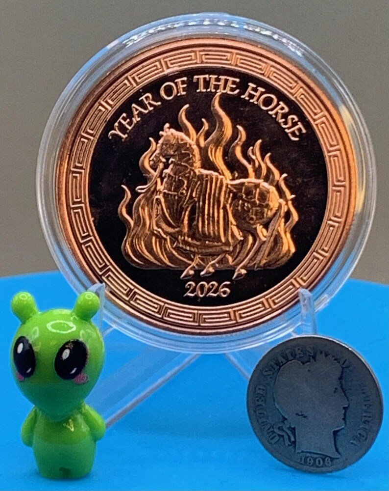 Encapsulated 2026 Year of the Horse copper round on a blue stand, armored horse obverse showing, flanked by the Area 51 Precious Metals green alien mascot figurine and a silver Barber dime for scale.
