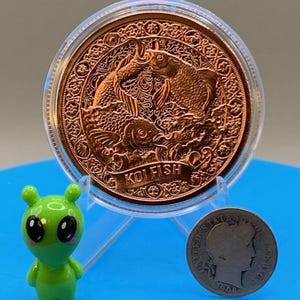 以下が含まれることがあります： 透明なプラスチック製保護ケースに入った、2匹の鯉が浮き彫りになった銅色の丸いコイン。コインには「KOI FISH」の文字が刻まれている。その横には、小さな緑色のエイリアンのフィギュアと、1906年製の銀色の米国コインが置かれている。