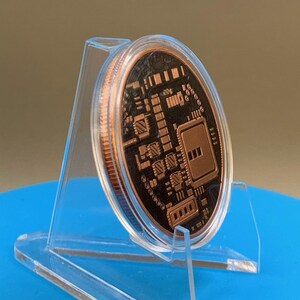 A circular copper-colored round featuring a black recessed surface with a detailed, raised circuit board pattern. The round is displayed in a clear, angled plastic stand, showing its metallic edge and intricate electronic design.