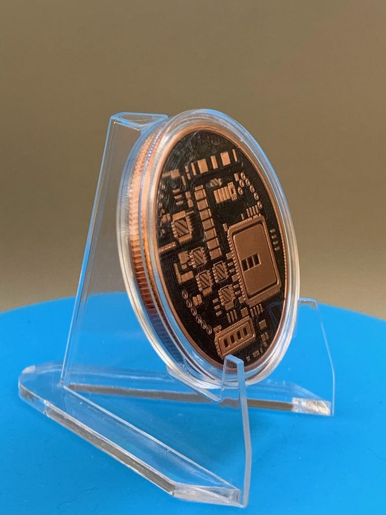 A circular copper-colored round featuring a black recessed surface with a detailed, raised circuit board pattern. The round is displayed in a clear, angled plastic stand, showing its metallic edge and intricate electronic design.