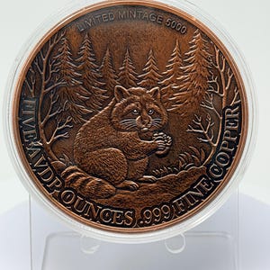 May include: A round copper coin featuring a raccoon in a forest setting. The text reads LIMITED MINTAGE 5000, FIVE AVD P OUNCES, and .999 FINE COPPER. The coin is encased in a clear protective plastic capsule and sits on a transparent display stand.