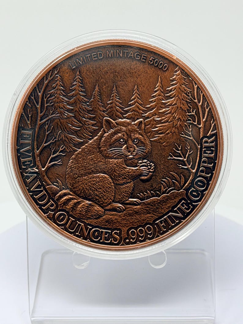May include: A round copper coin featuring a raccoon in a forest setting. The text reads LIMITED MINTAGE 5000, FIVE AVD P OUNCES, and .999 FINE COPPER. The coin is encased in a clear protective plastic capsule and sits on a transparent display stand.