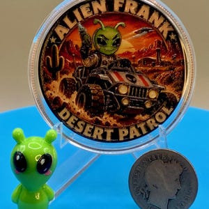 Alien Frank Desert Patrol 1 oz copper round in a clear capsule on a blue stand, shown next to a small green alien figurine and an older silver-colored coin to give a sense of scale.